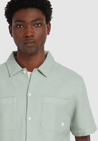 Lichtgroen shirt met korte mouwen van een gestructureerde stof, knoopsluiting aan de voorkant en twee opgestikte zakken; heeft een kraag en gestikte accenten.