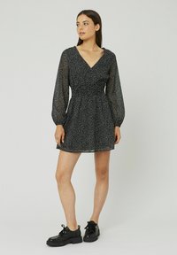 Robe noire avec de longues manches ballons transparentes, un décolleté en V, une taille smockée et des pois blancs. Portée avec des chaussures noires à lacets épaisses.