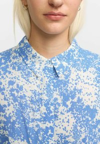 Light blue shirt with a scattered white speckle pattern. Features a classic collar and a single button at the neck. Smooth fabric texture.