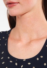 Námořnická halenka s kulatým výstřihem, zdobená malými zlatými listovými vzory. Tkanina se zdá být měkká s mírnou texturou. Okraj výstřihu je hladký.