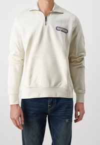 Pullover leggero in crema con zip a un quarto, tasca frontale, logo ricamato e polsini a costine. Tessuto morbido con design casual.