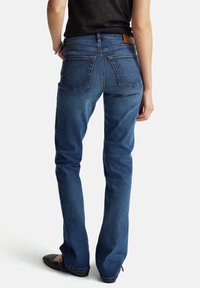 Jeans de mezclilla azul oscuro con una forma ajustada, bolsillos traseros visibles y un parche cerca de la cintura. Textura suave y diseño minimalista.