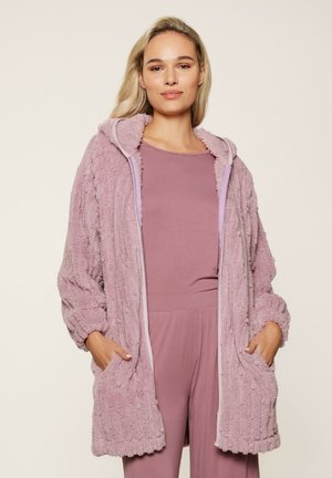 Robe en peluche violet avec un extérieur texturé, capuche, poches avant et bordures contrastantes. Porté sur un ensemble doux assorti de haut et bas.
