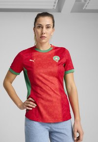 Röd fotbollströja med gröna detaljer, med geometriska mönster, rundad halsringning och Puma-logotyp på vänster bröst.