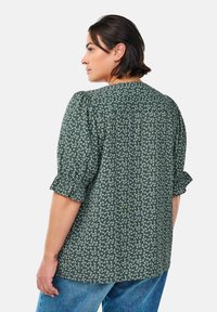 Blusa floreale verde con maniche a sbuffo, realizzata in un tessuto morbido, con scollatura rotonda e delicato motivo di fiori bianchi su uno sfondo scuro.