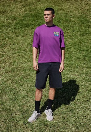 Egy férfi áll a fűben, lila "Venice Beach Hip Hop" pólót, fekete rövidnadrágot, fekete zoknit és fehér tornacipőt visel, balra néz.
