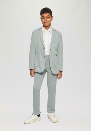 Hellgrauer Anzug mit zwei Taschen, ein gefütterter Blazer und passende Hose, kombiniert mit einem weißen Hemd mit kleinen blauen Mustern und weißen Turnschuhen.