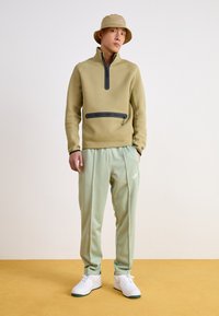 Μπεζ φλις μπλούζα με φερμουάρ και μαύρη τσέπη μπροστά, σε συνδυασμό με ανοιχτό πράσινο παντελόνι φόρμας. Λευκά αθλητικά παπούτσια με πράσινες λεπτομέρειες ολοκληρώνουν την εμφάνιση.