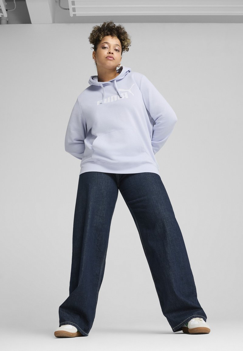 Jasnofioletowa bluza z kapturem z kieszenią kangurką z przodu oraz białym logo, zestawiona z szerokimi ciemnoniebieskimi dżinsami i białymi trampkami.