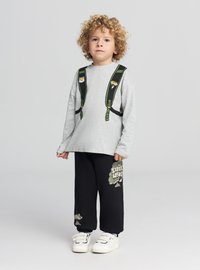 Maglietta grigia a maniche lunghe con stampa di uno zaino, abbinata a pantaloni della tuta neri con grafica e testo verdi, completati da sneakers bianche.