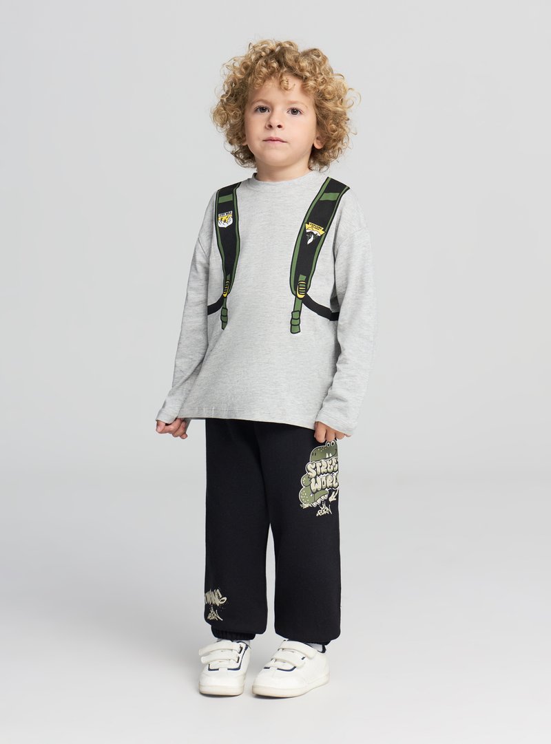 Maglietta grigia a maniche lunghe con stampa di uno zaino, abbinata a pantaloni della tuta neri con grafica e testo verdi, completati da sneakers bianche.