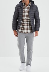 Giacca imbottita nera con cappuccio, camicia a quadri marrone, crema e grigia, pantaloni della tuta grigio chiaro e scarpe da ginnastica bianche. Outfit casual.