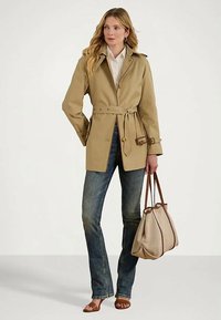 Nainen, joka on pukeutunut beigeen vyötettyyn trench-coattiin, valkoiseen paitaan, sinisiin farkkuihin ja ruskeisiin sandaaleihin, pitää suurta beigeä käsilaukkua, jossa on ruskeat hihnat.