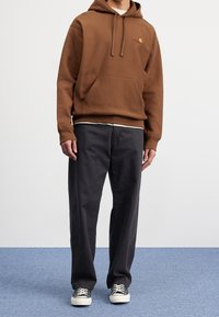 Felpa marrone con cappuccio, tasca a marsupio, cordoncino e piccolo logo. Abbinata a pantaloni neri larghi e scarpe da ginnastica nere con suola bianca.