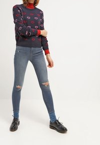 Maglione navy con collo a coste rosso, caratterizzato da motivi a cuore rosa e bianchi. Jeans skinny strappati blu chiaro, abbinati a scarpe nere con lacci.