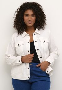 Chaqueta de mezclilla blanca con dos bolsillos en el pecho, detalles en botón negro y un corte relajado, combinada con una blusa negra y jeans azules.
