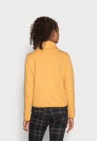 Polo de forro polar amarillo con cuello alto, textura suave y corte relajado. Combinado con pantalones de cuadros que presentan tonos oscuros y claros.