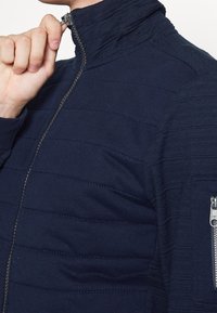 Chaqueta de cremallera azul marino con textura acolchada, cuello alto, bolsillo lateral con cremallera y costuras contrastantes.