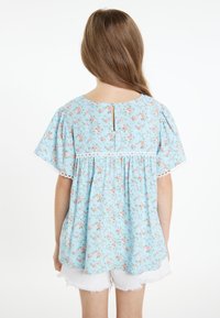 Hellblaues Blumenshirt mit kurzen Ärmeln, ausgestattet mit weißen Spitzenbesätzen und einem Schlüsselloch-Rücken. Hergestellt aus leichtem Stoff. Getragen über weißen Shorts.