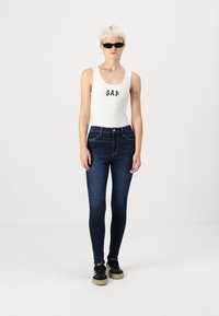 Fehér, testhezálló atlétatrikó "GAP" logóval, sötétkék skinny farmernadrág és fekete sportcipő, egy sima fehér háttér előtt állva.