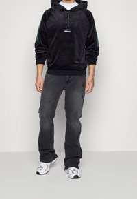 Hoodie in velluto nero con righe verdi laterali, colletto con mezzi zip e dettaglio del logo; abbinato a jeans grigio scuro e scarpe da ginnastica bianche.
