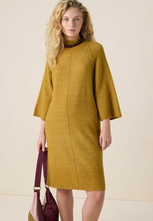 Femme aux longs cheveux blonds ondulés, portant une robe en maille jaune moutarde et un collier de perles foncé, tenant un sac à main bordeaux et rose.