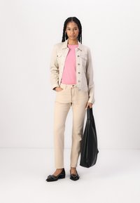 Beige cotton jacket over a pink t-shirt, paired with beige striped trousers and black loafers. Holding a black tote bag.