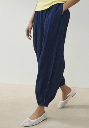 Person iført løstsiddende navyblå teksturerede bukser med elastiske manchetter, en lys gul top og hvide mesh slip-on sko, stående på en lys gulv.