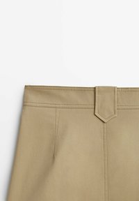 Partie supérieure d’un pantalon beige montrant la ceinture avec détail de boucle et couture double visible sur tissu lisse.