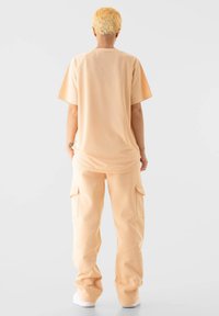 Trumpomis rankovėmis, lengvas, tinklelio megztinis šviesiai persikinės spalvos, derinamas su krovinių stiliaus kelnėmis tokios pat spalvos; abu drabužiai turi laisvą kirpimą.