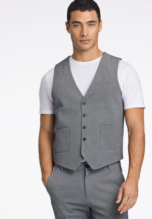 Grahamfarbene Weste mit V-Ausschnitt-Design, ausgestattet mit fünf Knöpfen, zwei vorderen Taschen und einer klaren, maßgeschneiderten Silhouette. Getragen über einem weißen Hemd.