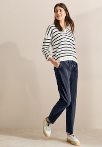 Weiße und marineblaue gestreifte Strickjacke mit V-Ausschnitt und dreiviertellangen Ärmeln, kombiniert mit dunkelblauen Zugband-Hosen und weißen Sneakers mit beigen Sohlen.