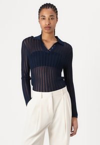 Navy blue, ribbed knit polo shirt with a collar, featuring a sheer design and button placket. Worn with high-waisted cream trousers.