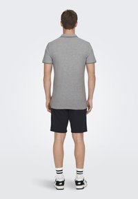Graues Poloshirt mit kurzen Ärmeln im Standardfit, schwarze Besätze an den Ärmeln, kombiniert mit marineblauen Shorts und weißen/schwarzen Sportschuhen.