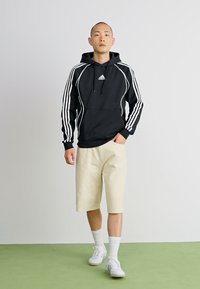Sweatshirt preto com capuz e riscas brancas, calções bege e ténis brancos. O material é suave, com mangas de comprimento médio e punhos elásticos.