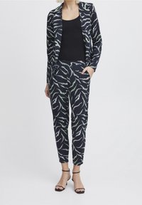 Žena v černém tílku se shodným černým sakem a kalhotami s bílým a zeleným listovým vzorem, doplněná o černé sandály na podpatku.