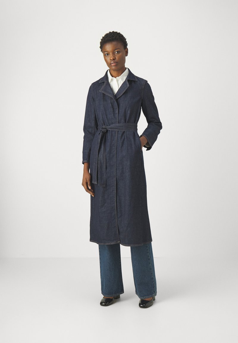 Cappotto trench in denim, blu scuro, di lunghezza media, con cintura in vita, colletto a scollatura a punta, maniche lunghe, tasche laterali e cuciture a contrasto.