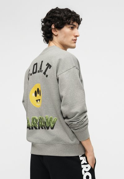 Νεαρός άνδρας με κυκλικά μαλλιά που φοράει γκρι φούτερ με το κείμενο "G.O.A.T" και κίτρινο γραφικό χαμογελαστό πρόσωπο στην πλάτη, μαύρο παντελόνι με λευκούς χαρακτήρες.