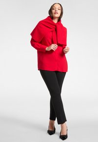 Czerwona koszula na guziki z długim rękawem w połączeniu z czarnymi dopasowanymi spodniami i czarnymi czółenkami na pasku z tyłu, uzupełniona czerwonym szalem narzuconym na ramiona.