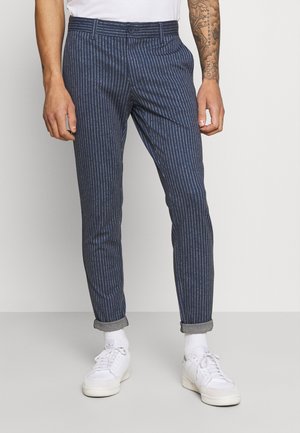 Pantalon rayé bleu marine à coupe ajustée, avec une fermeture à bouton, des ourlets retroussés et un tissu léger. Porté avec des baskets blanches.