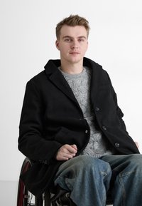 Schwarzer Wollmantel mit Reverskragen, Knopfverschluss und seitlichen Taschen über einem grauen Zopfstrickpullover; Modell sitzt im Rollstuhl.