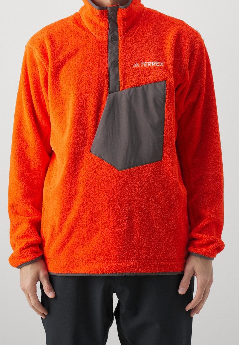Oranje fleece pullover met een hoge kraag, voorzien van een donkerbruine borstzak en knoopdetails aan de voorkant. Zachte textuur overal.