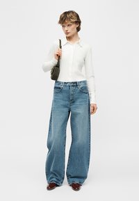 Woman wearing a white button-up top, wide-leg blue jeans, brown shoes, and carrying a small black shoulder bag, standing against a plain background.