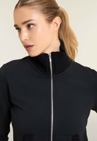 Chaqueta negra con cremallera que presenta un cuello acanalado, textura suave y dos bolsillos delanteros con un diseño estructurado. Detalle de cremallera metálica sutil.