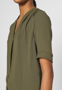 Torso superiore di una donna che indossa una blusa verde oliva con scollo a V e maniche corte arrotolate, su uno sfondo grigio uniforme.