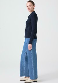 Mörkblå randig cardigan med knäppning fram, tillsammans med vida ljusblå jeans med splitt söm och fransad fåll. Klassisk skodon kompletterar outfitten.