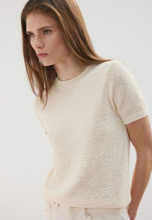 Młoda kobieta z długimi brązowymi włosami, ubrana w kremowy sweter z krótkim rękawem i jasne spodnie, patrząca lekko na bok.