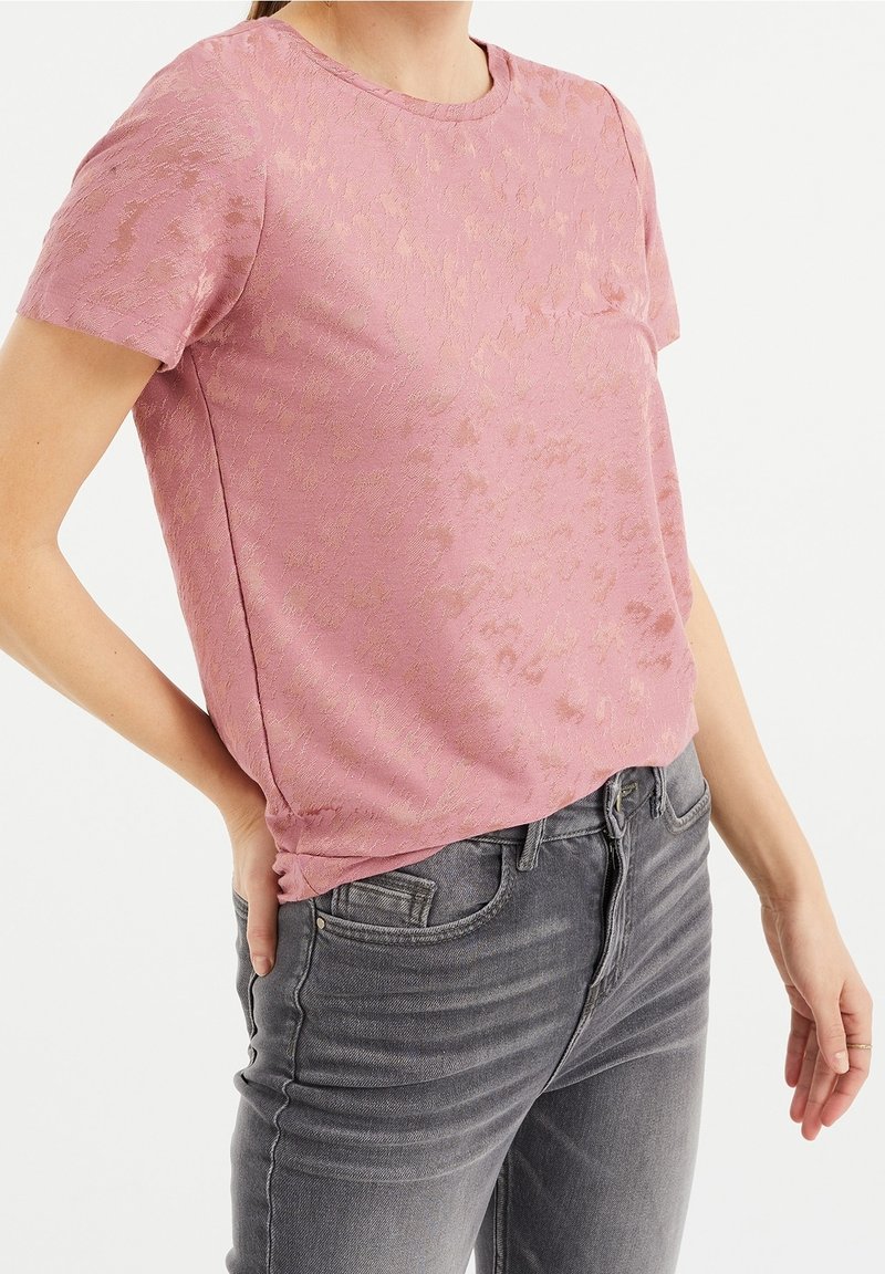 Krátký rukáv, světle růžový top s jemným texturovaným vzorem, kulatým výstřihem a bočními švy, spárovaný se šedými džíny.