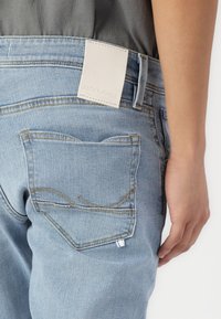 Lyseblå denimjeans har en jevn tekstur, subtile sømmer på baklommene, og en hvit lærmerkelapp i livet.