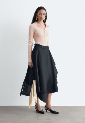 Femme portant un haut rose clair à manches longues, une jupe asymétrique noire, des talons slingback noirs, des boucles d'oreilles dorées, tenant un sac noir avec un foulard jaune.